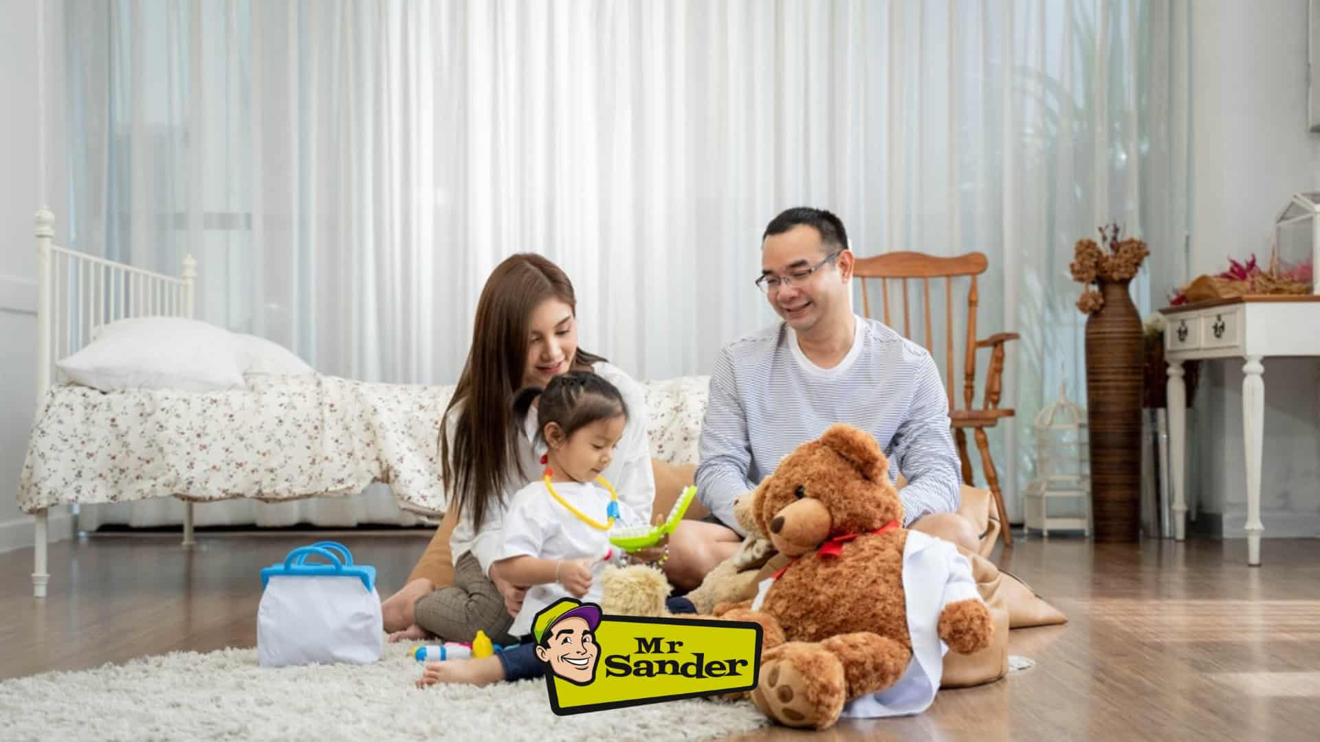 A family sits on a plush rug in their cozy living room in Stoke Newington, UK, enjoying the beautifully sanded and refinished oak hardwood flooring done by Mr Sander®. The room features light, airy curtains, a white bed with floral bedding, and charming furniture, showcasing the high-quality restoration work by Mr Sander®.