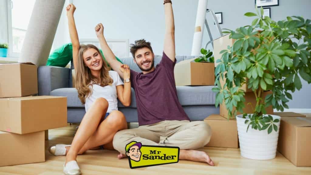 A joyful couple sitting on newly sanded and refinished light oak hardwood flooring, celebrating their new home in Farnborough, UK. The room is filled with moving boxes, a rolled-up carpet, and a potted plant, showcasing the beautiful work done by Mr Sander®.