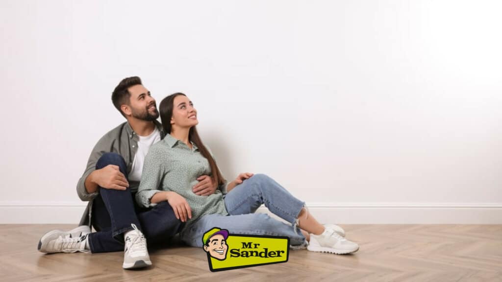 A couple sitting on the floor, admiring their newly refinished herringbone pattern oak flooring. The floor's detailed craftsmanship and smooth finish create a warm and inviting atmosphere.