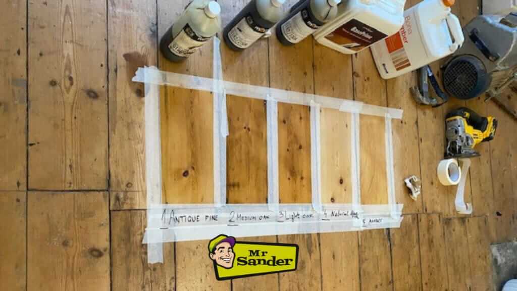 Wooden floor with sample finishes labeled Antique Pine, Medium Oak, Light Oak, Natural Oak, and Amber, alongside various finishing products and tools.