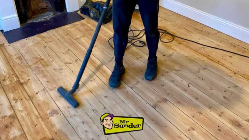 A professional worker using a vacuum cleaner to clean up dust and debris from a freshly sanded wooden floor. The floor has a smooth and even finish, showcasing the results of the sanding process. The Mr Sander® logo is visible in the lower center of the image.