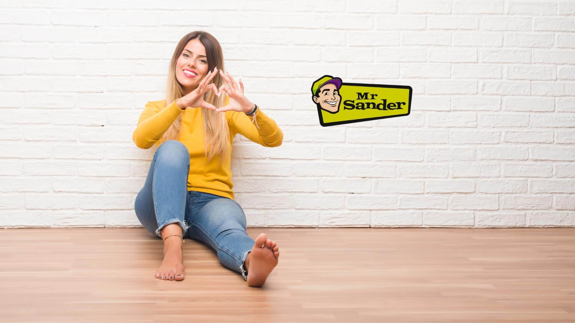 A woman sitting on a beautifully refinished floor in Elephant & Castle, UK, done by Mr Sander®. She is wearing a yellow sweater and blue jeans, smiling and forming a heart shape with her hands against a white brick wall. The Mr Sander® logo is visible on the wall.