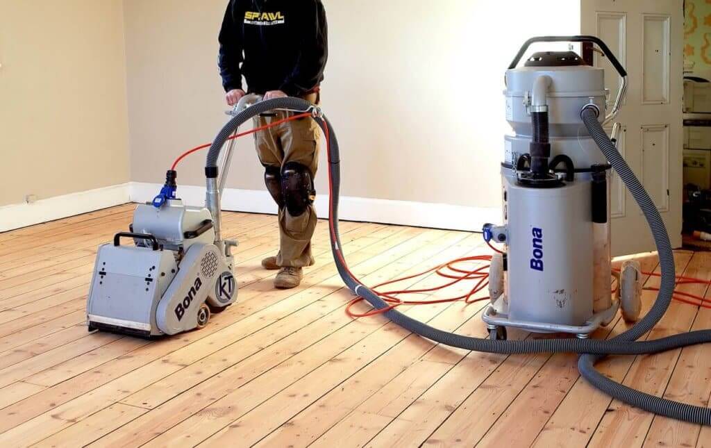 A professional technician sanding a wooden floor using a Bona floor sander connected to a dust collection system for a clean and efficient process.
