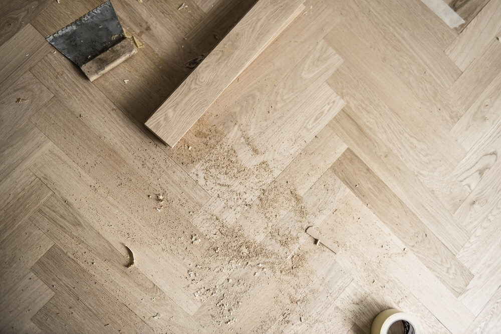 Top-down view of a light-colored hardwood floor with a herringbone pattern being sanded and refinished. Tools like a trowel and measuring tape are scattered around the work area.