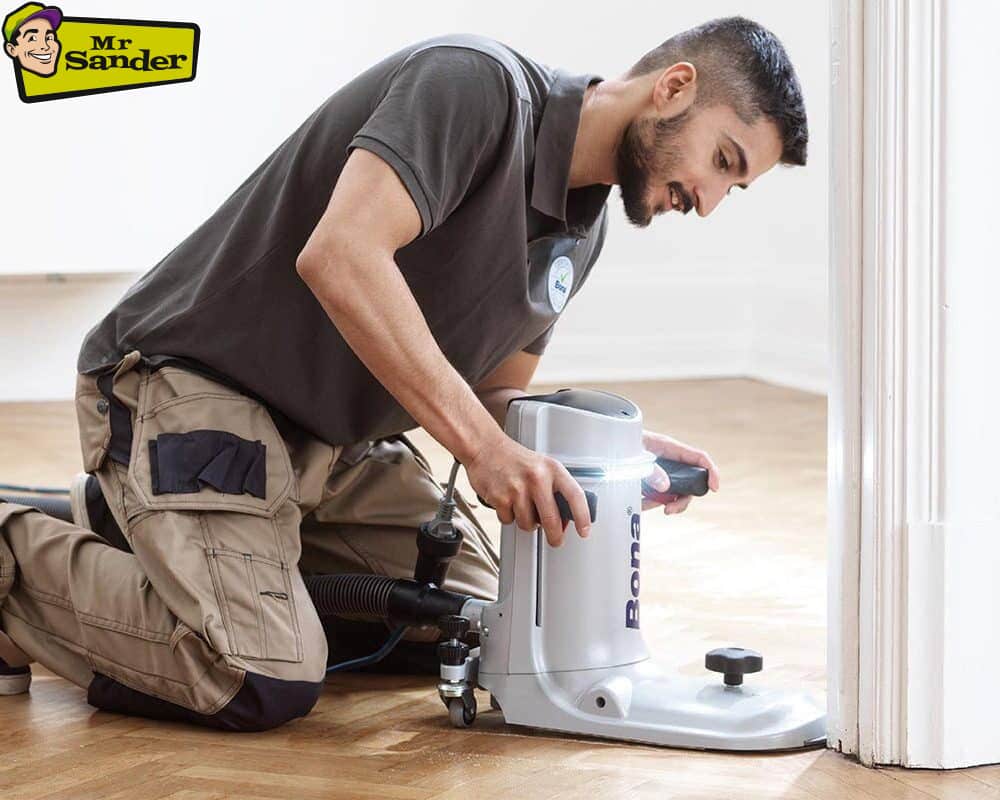 A professional floor sanding technician kneels while using a specialized edge sander to work on a herringbone wooden floor. The Mr Sander® logo is displayed in the top-left corner.
