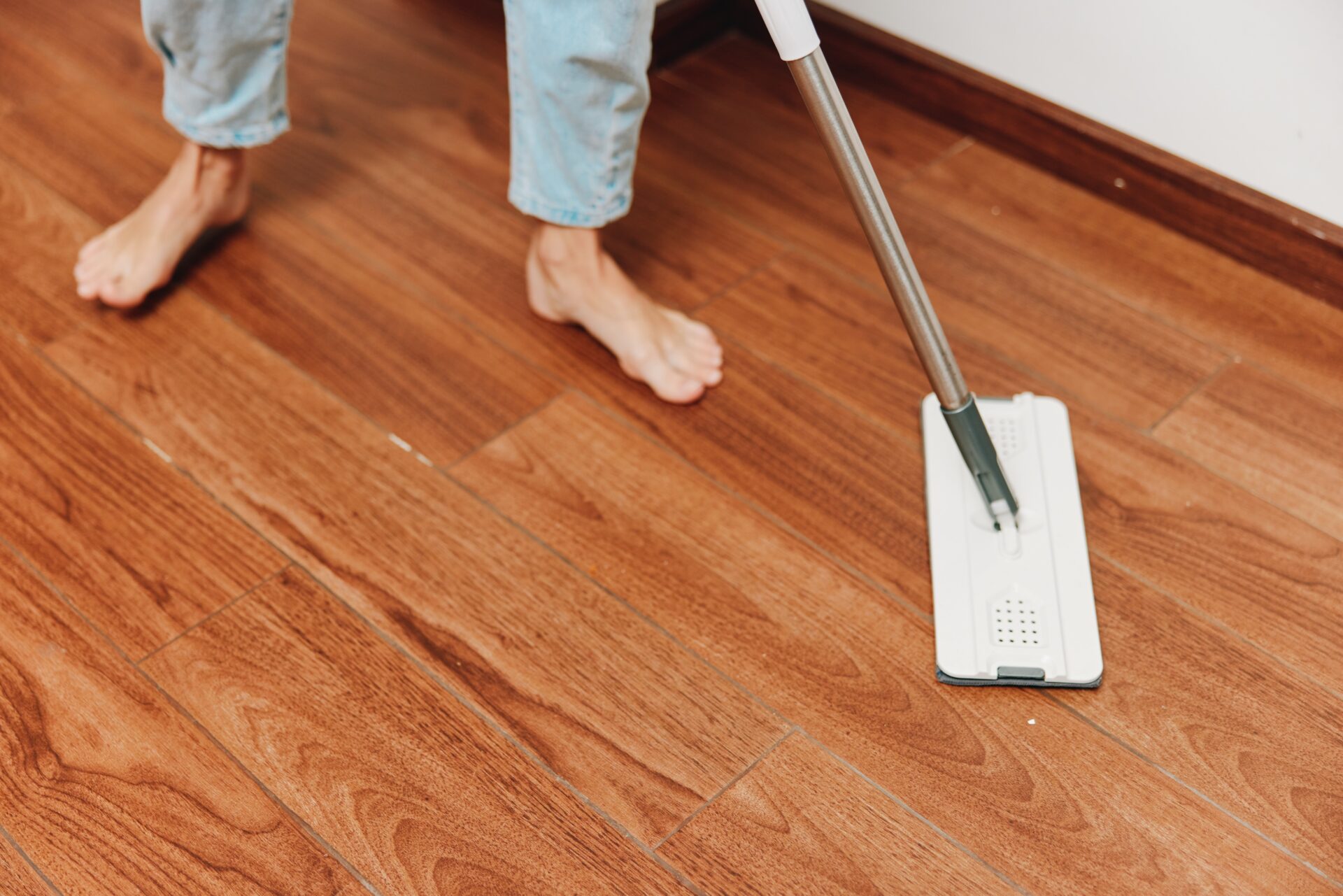 Bare-foot homeowner using a flat mop to maintain a freshly completed hardwood floor installation with warm chestnut tones.