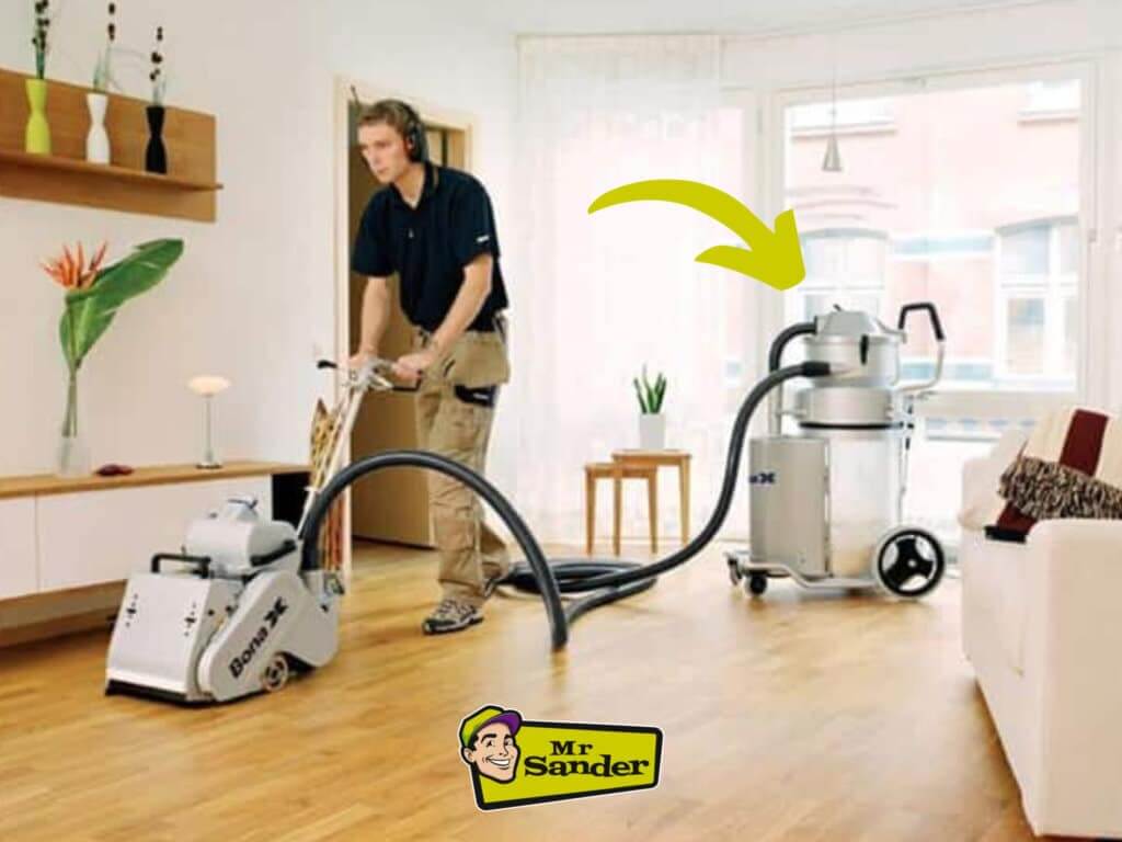 A professional using a floor sanding machine equipped with an advanced dust collection system in a bright, modern living room. The sanding machine connects to a large vacuum canister with hoses, effectively capturing dust to maintain a clean and safe workspace. The "Mr Sander" logo is featured in the bottom-right corner.