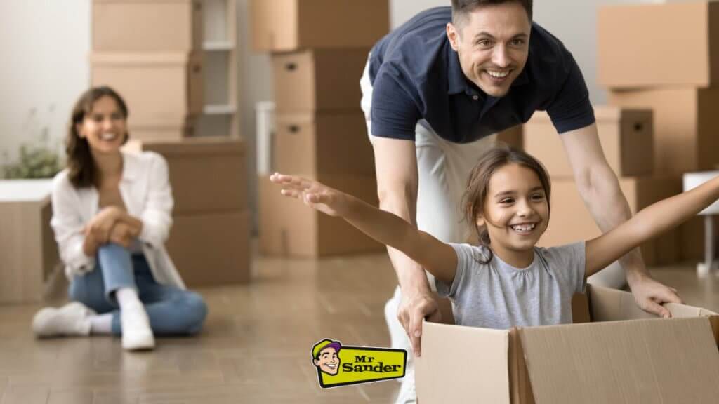A smiling child seated in a cardboard box with arms outstretched, being pushed by an adult while another adult looks on amidst moving boxes in a bright room.