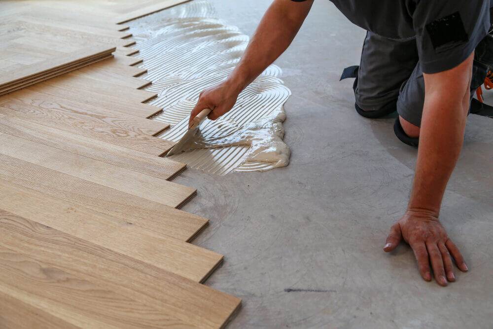 Flooring installer spreading adhesive on a concrete subfloor to secure herringbone planks during a Parquet Floor Fitting