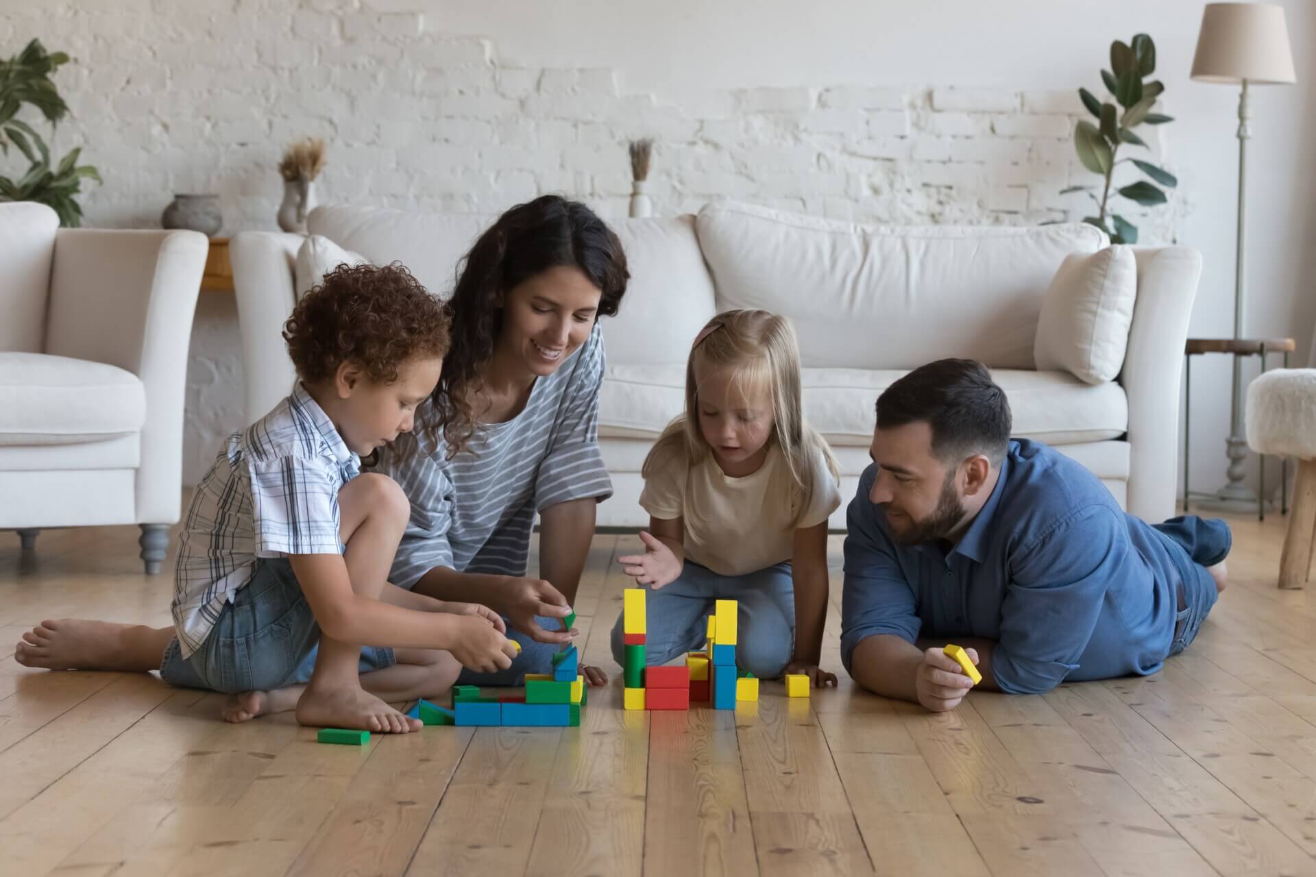 A smiling family of four, including two young children, playing with colourful building blocks on a newly installed wood floor.