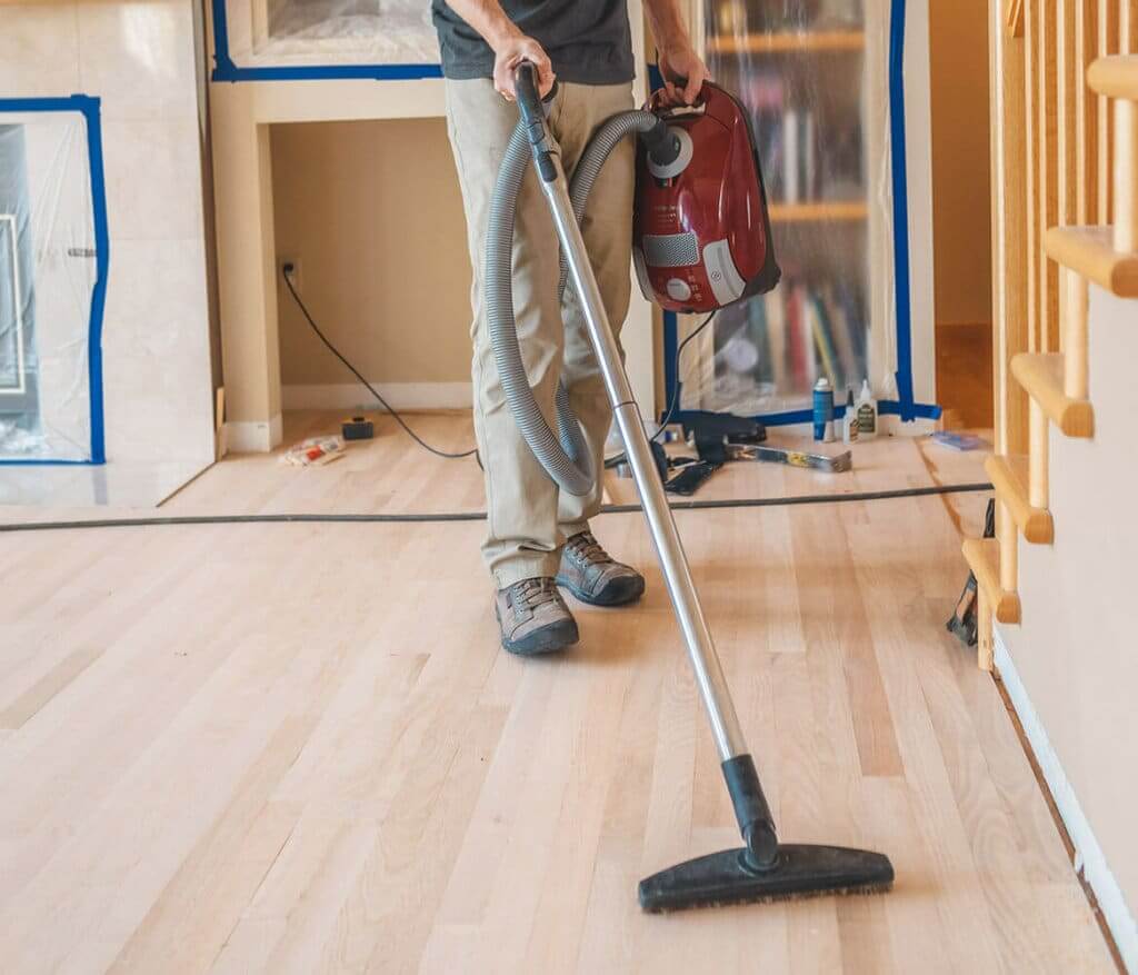 A professional from Mr Sander® vacuuming dust from a recently sanded wooden floor in a living area. The room is prepared for refinishing with plastic coverings on the furniture and tape on the walls. The professional uses a red canister vacuum to ensure a clean surface before applying the finish.