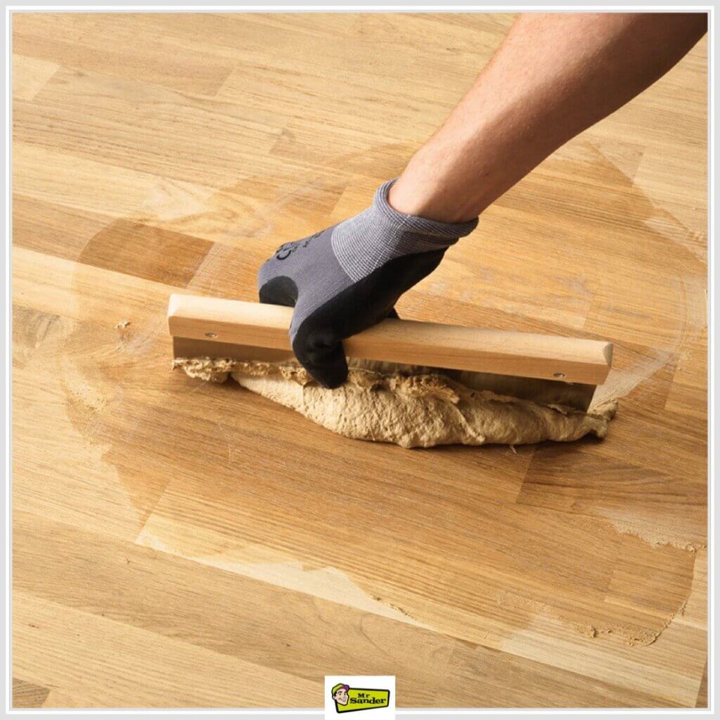 A person wearing a glove uses a wooden trowel to spread a resin-based filler over hardwood floor gaps.