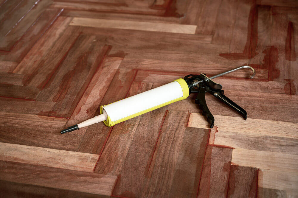 A caulk gun resting on a newly installed parquet floor with red adhesive lines, illustrating the final sealing process in a West London Floor Fitting project.