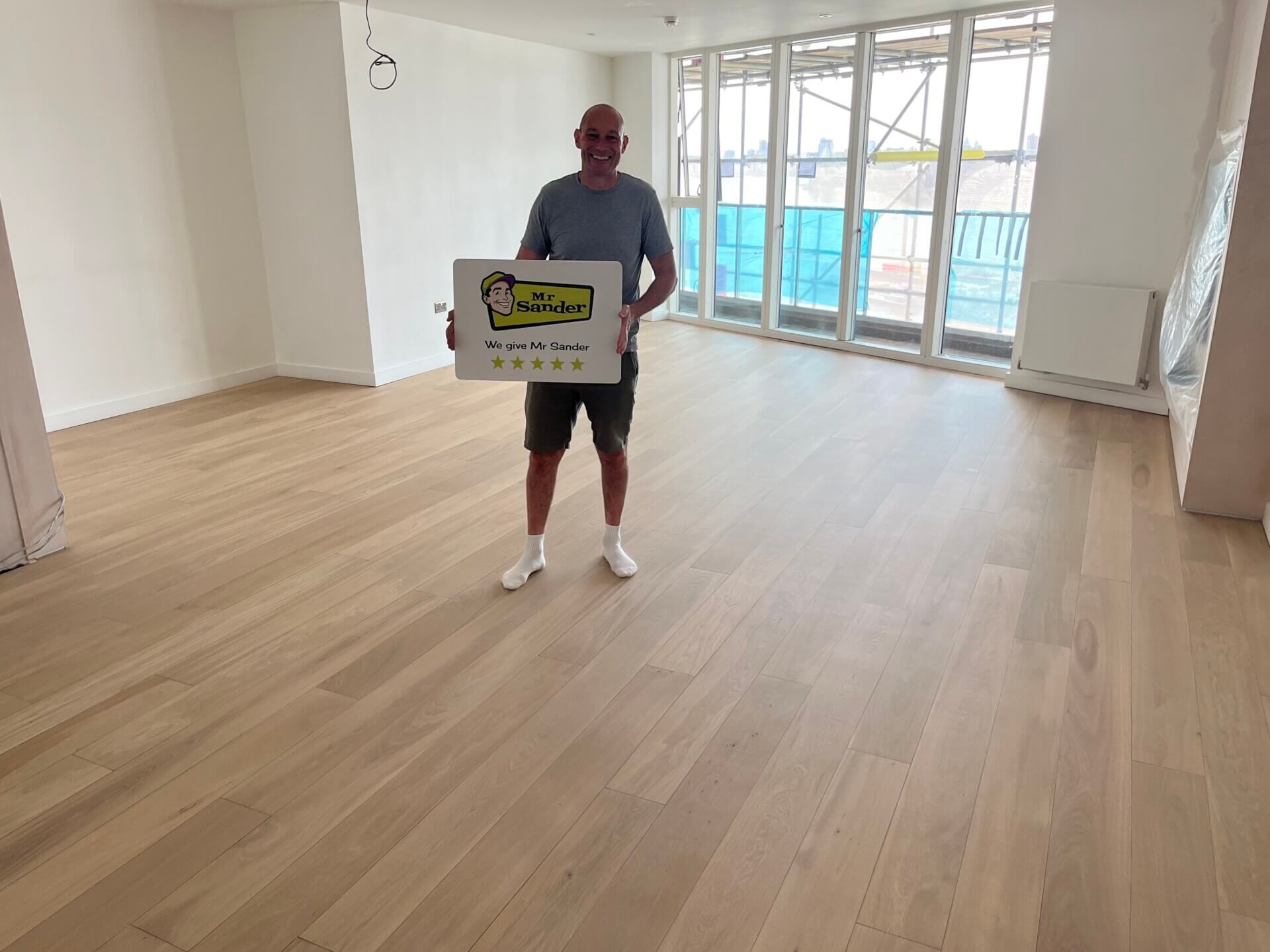 A satisfied Mr Sander customer in London standing on newly installed engineered flooring, holding a sign with five-star feedback
