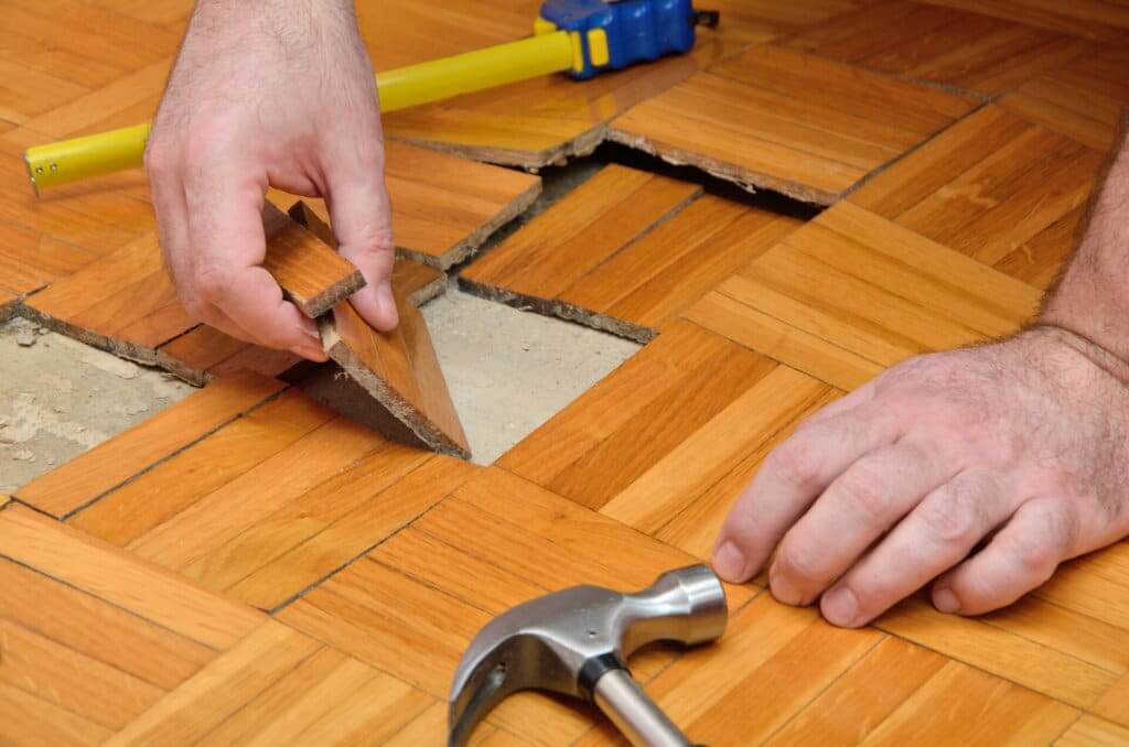 A close-up of a person replacing damaged parquet tiles on a wooden floor. The individual is carefully fitting a new tile into the space, with a hammer and tape measure visible nearby, indicating a repair in progress.