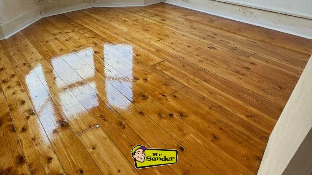 A beautifully restored wooden floor with a high-gloss finish, reflecting natural light from a nearby window, highlighting the rich grain and warm tones of the wood.