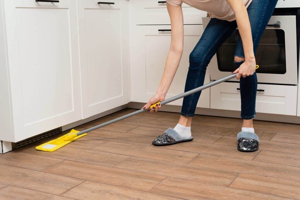 A person is cleaning a kitchen floor with a yellow mop, reaching under white cabinetry. The individual is wearing casual clothing, including jeans and cozy slippers, while bending over to ensure a thorough cleaning. The floor has a wooden appearance, and the kitchen has modern white cabinets and appliances.