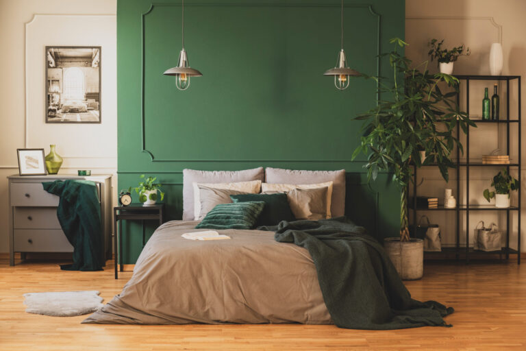 Bedroom-style greenhouse setting with Natural Oak solid strip wood flooring, deep green feature wall, potted money tree and indoor plants around a grey-linen bed.