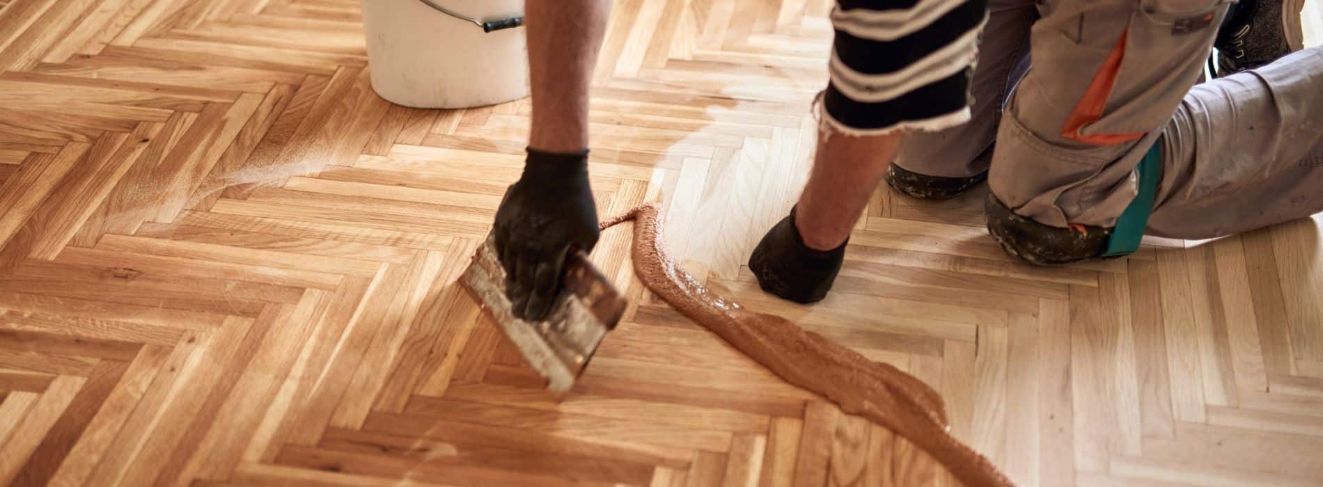  A close-up photograph of a person using a brush on a wooden floor. The individual is likely in the process of applying a finishing coat, such as varnish or sealant, to the wooden surface. The brush strokes can be seen in detail, showcasing the precision and care required to achieve a smooth and even finish. The wood's natural grain and color are visible, providing an overall warm and inviting aesthetic. The image conveys the intricacies of the flooring process, highlighting the attention to detail and expertise required for achieving beautiful and lasting results.