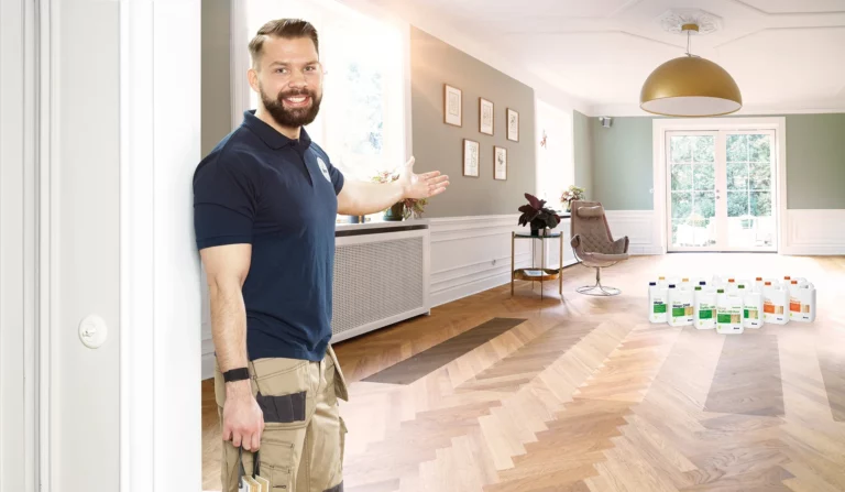 A smiling floor sanding professional presenting a beautifully refinished herringbone wood floor treated with eco-friendly finishes, with several Bona Eco-Friendly Floor Finishes containers displayed in the background.