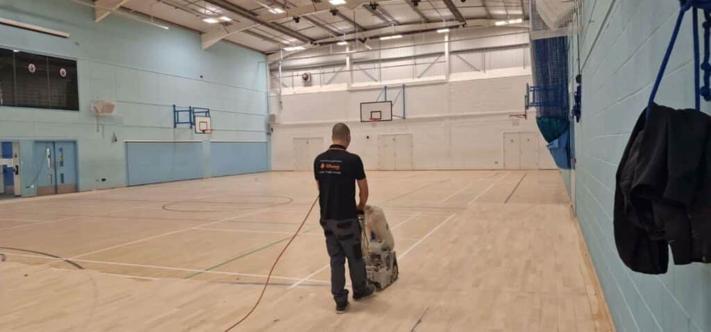 A man is operating a floor sanding machine in an indoor basketball court. He is walking away from the camera, focused on sanding the wooden floor. The gym is spacious and well-lit with fluorescent lights, and there are basketball hoops mounted on the walls. The walls are painted in a light blue color, and there is sports equipment stored along the wall. The environment suggests maintenance work in progress in a community or school sports facility.