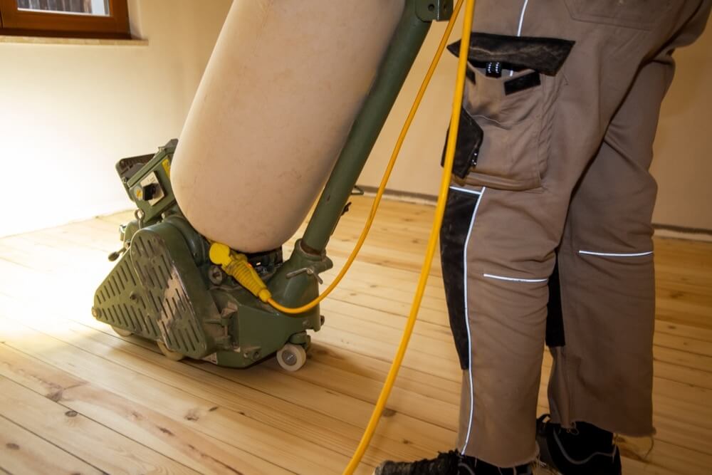 Worker operating a floor sanding machine on a pine wood floor in a home located in, UK. The image shows the process of sanding to restore the floor before refinishing, with the worker standing in the corner of the room.