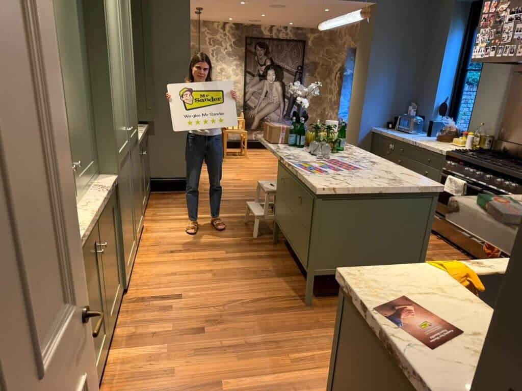 A person standing in a beautifully restored kitchen with polished wooden flooring, holding a sign that says "Mr. Sander - We give Mr. Sander 5 stars".