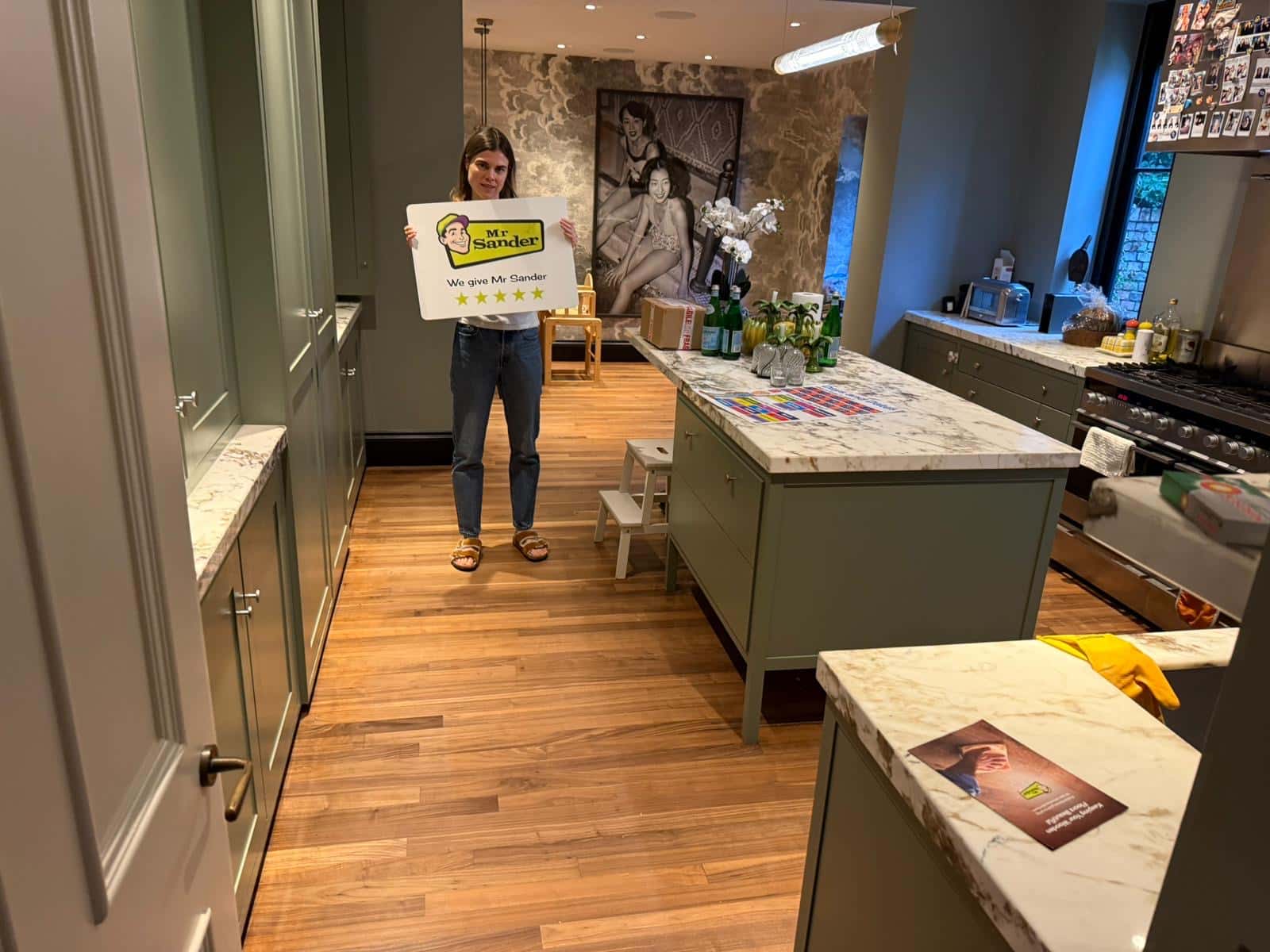 A person standing in a beautifully restored kitchen with polished wooden flooring, holding a sign that says "Mr. Sander - We give Mr. Sander 5 stars".