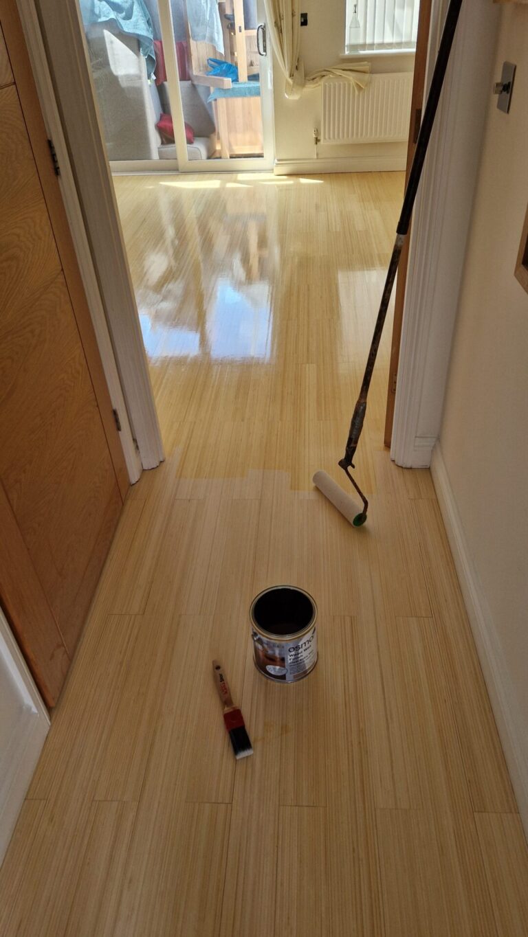 A newly refinished natural bamboo floor with a glossy finish, viewed from the hallway, where a paint roller, brush, and finish can are visible during the application process of professional floor sanding for bamboo floors.