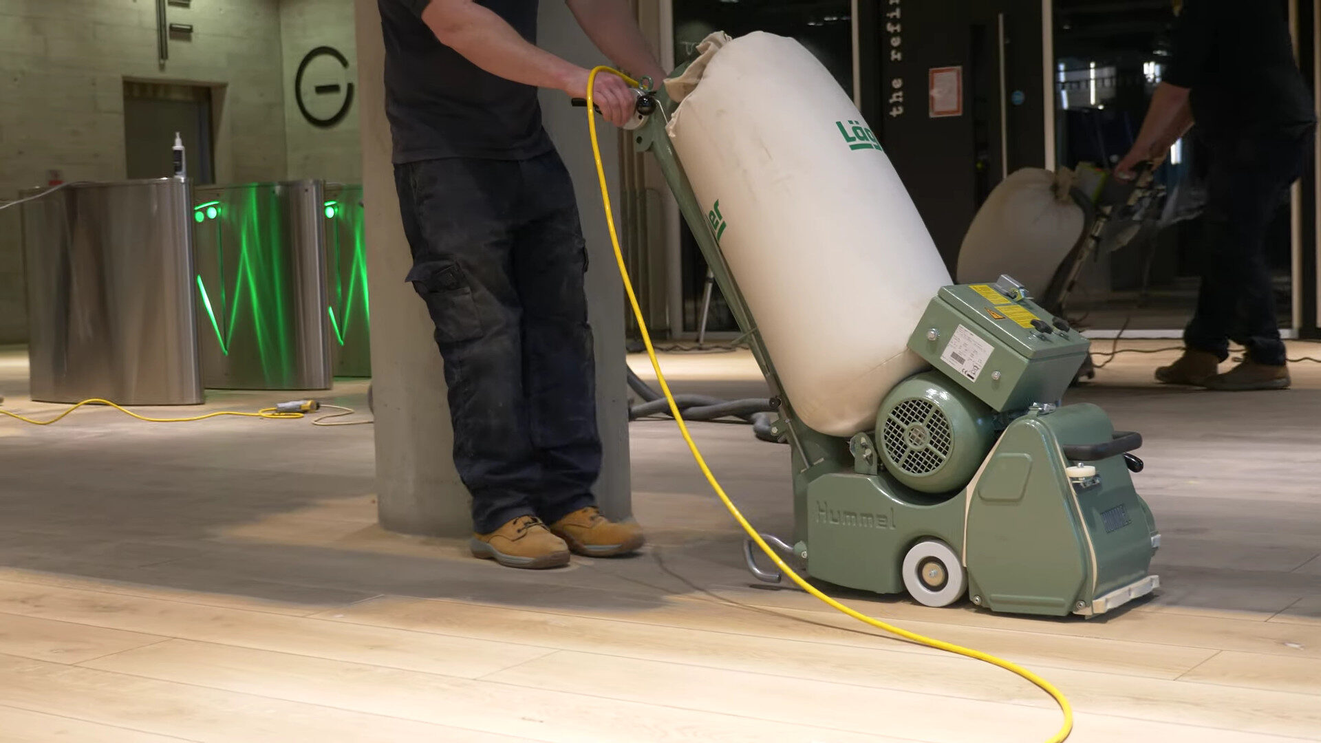 A flooring specialist operating a commercial-grade floor sander on a large wooden floor in a busy commercial setting.