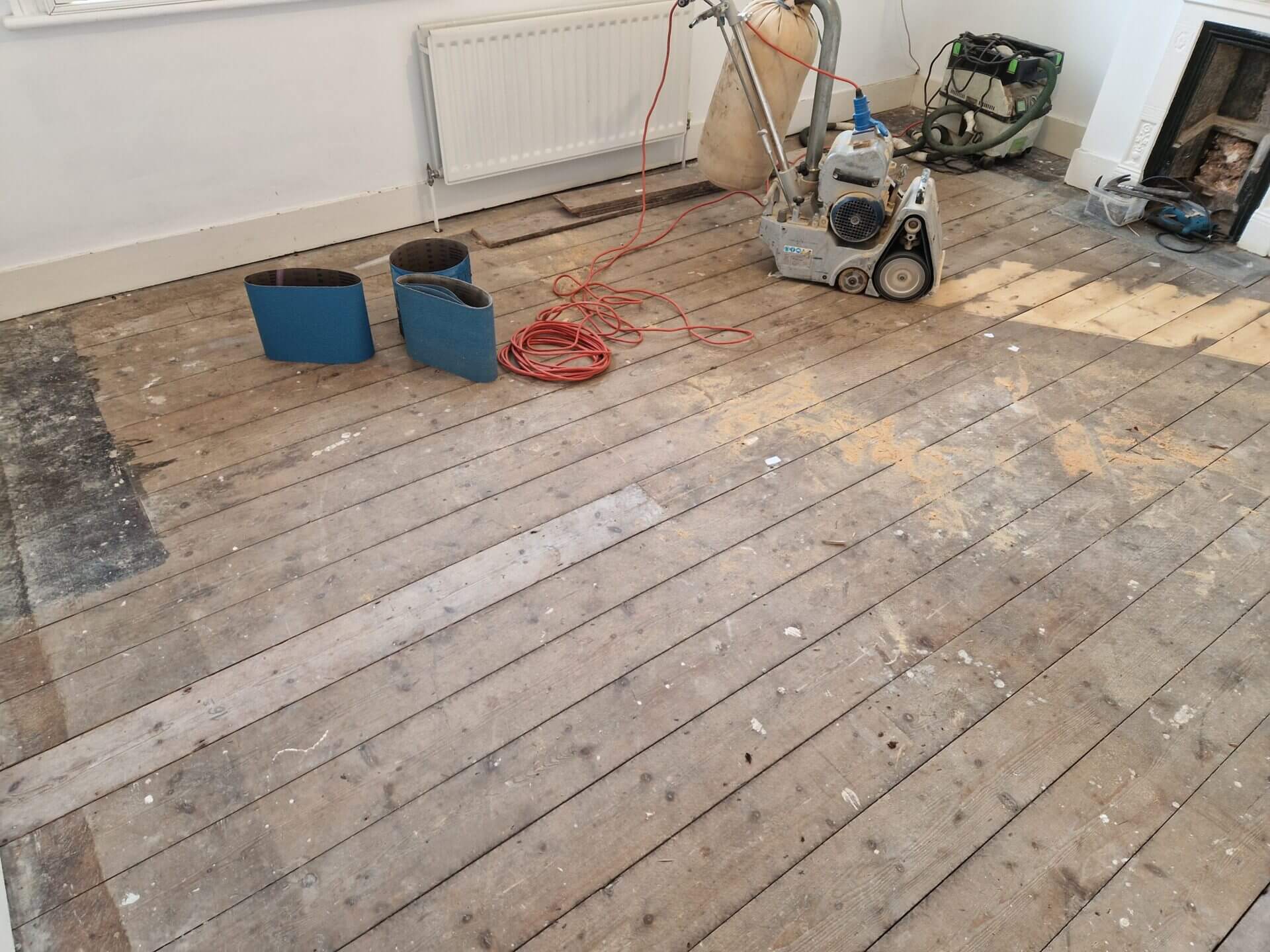 Partially sanded pine floorboards, sanding belts, and a belt sander in a living room, showcasing the initial stages of a Floor Sanding for Pine Floors project.
