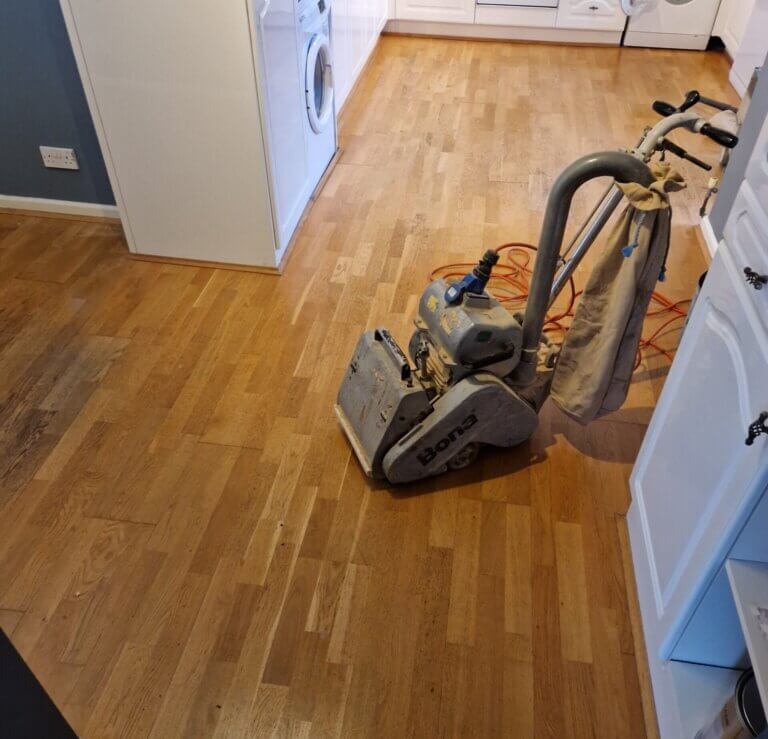 A Bona floor sander being used to restore a worn wooden kitchen floor, with visible dust bag and cables.