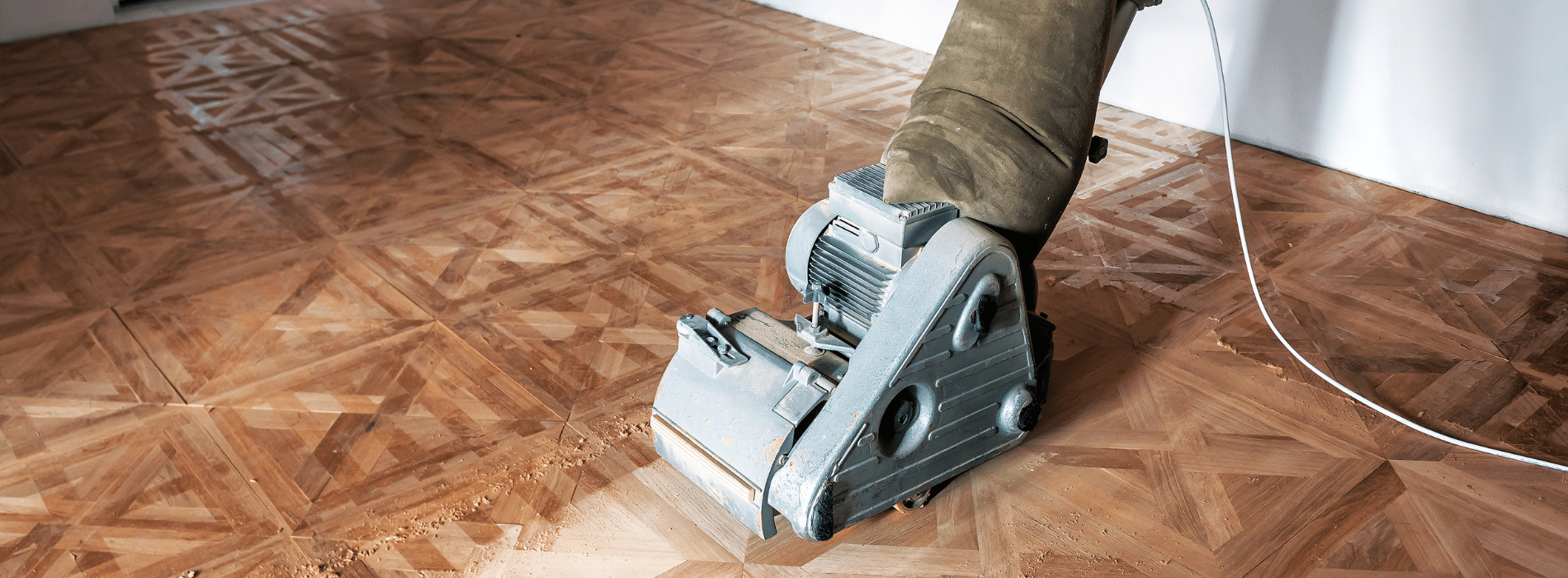 A skilled worker operating a Bona belt drum sander 240v, dimension 200mm on a herringbone floor in Hampstead, NW3. The worker's hands firmly grip the sander as they guide it across the surface. The sander is connected to a dust extraction system, effectively capturing the generated dust.