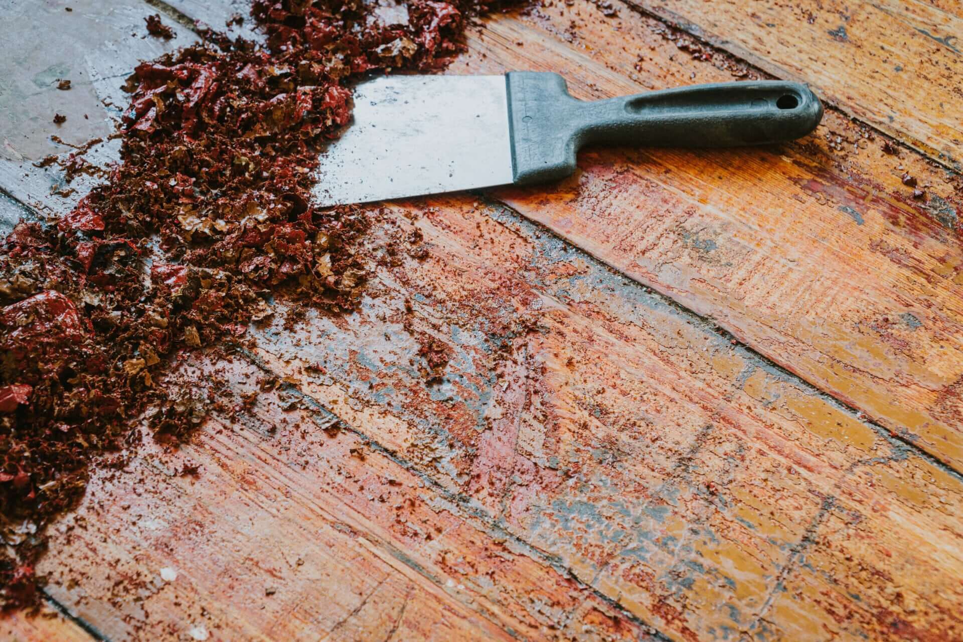 A black-handled scraper removing layers of red paint and finish from a worn wooden floor surface.