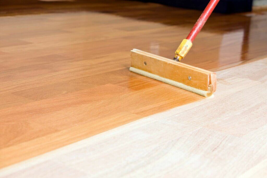 A close-up of a wooden floor being finished with a clear coating using a floor applicator tool. The image shows a clear contrast between the unfinished light wood and the newly coated section, which has a rich, glossy appearance. The process highlights the transformation during the floor refinishing.