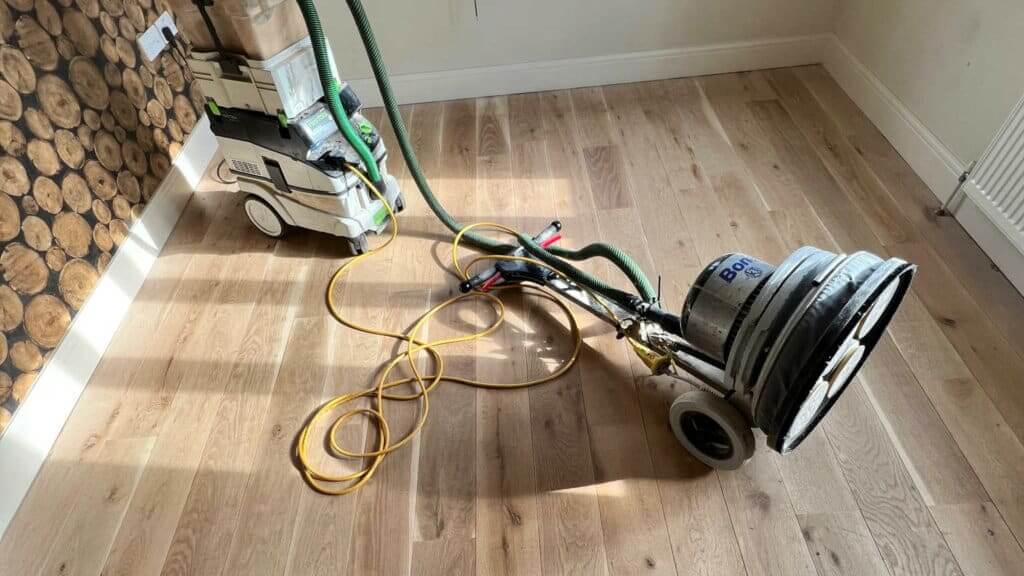 A man from Mr Sander® is sanding an oak floor in a living room in Catford, UK using a Bona FlexiSand 1.9 (AMO310002) buffer. The machine features a 1.9 kW motor, operates at 230 V and 50 Hz, with a disc diameter of 407 mm (16”). It has a rated current of 11 A, includes a 16 A overload switch, thermal protection, and dust containment system. The abrasive disc speed (unloaded) is 147 rpm, and the machine's total weight is 50 kg. Dimensions are 1170 mm height, 490 mm width, and 640 mm depth.