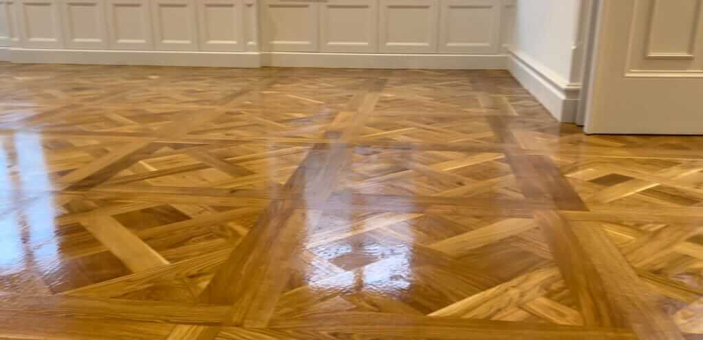 A beautifully restored parquet floor with a glossy, polished finish reflecting light. The intricate wooden pattern is vibrant and restored to its original richness, highlighting the transformation after sanding and refinishing. White paneling and molding along the walls complement the elegance of the restored floor.