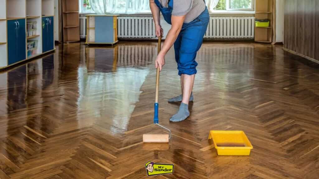 A floor refinisher using a roller to apply a shiny protective coat on a herringbone wood floor in a bright room.