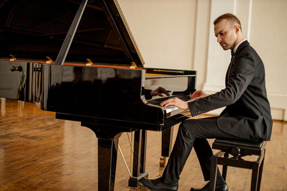 A man in a black suit playing a glossy black grand piano on a polished wooden floor.