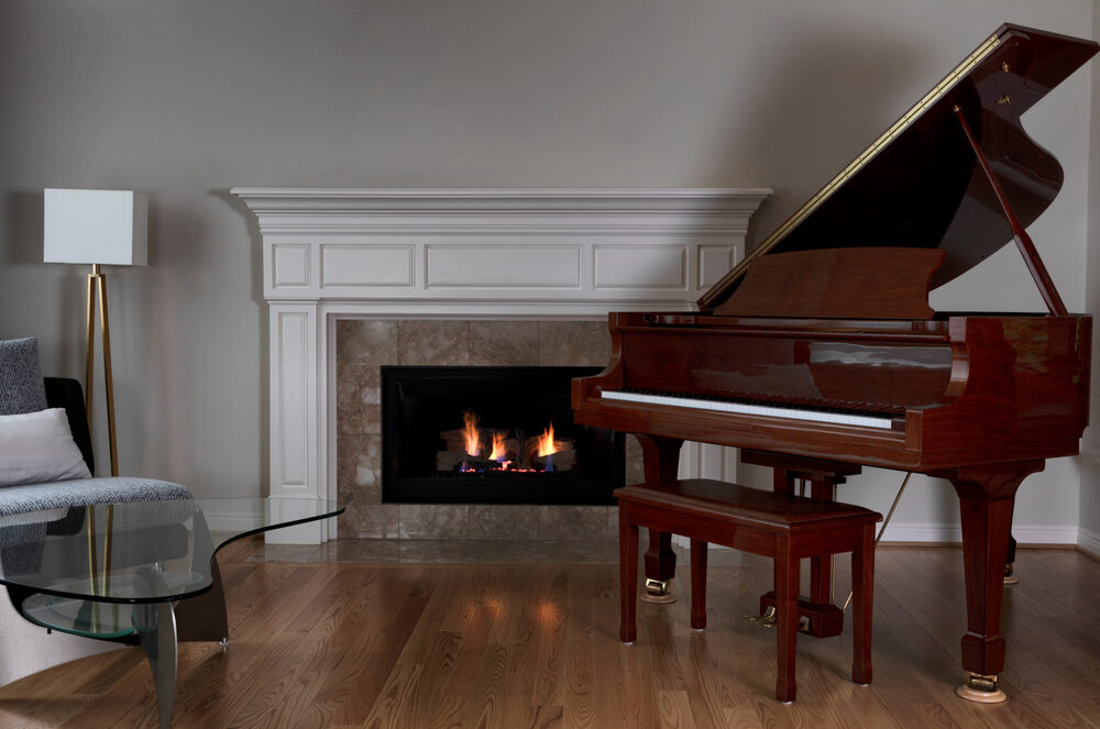A polished wooden grand piano positioned in a living room with a lit fireplace, glass coffee table, and hardwood floors.