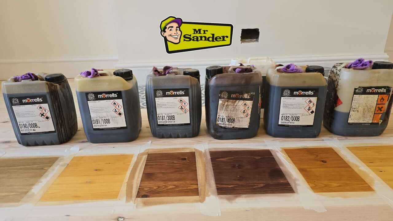 A lineup of Morrells brand wood finishing containers with various colour swatches on pine floorboards, showcasing eco-friendly stains and sealants for modern floor sanding projects.