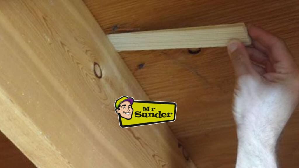 A hand is seen inserting a wooden shim into a gap in a pine floorboard, demonstrating a repair technique on a refinished wooden floor.
