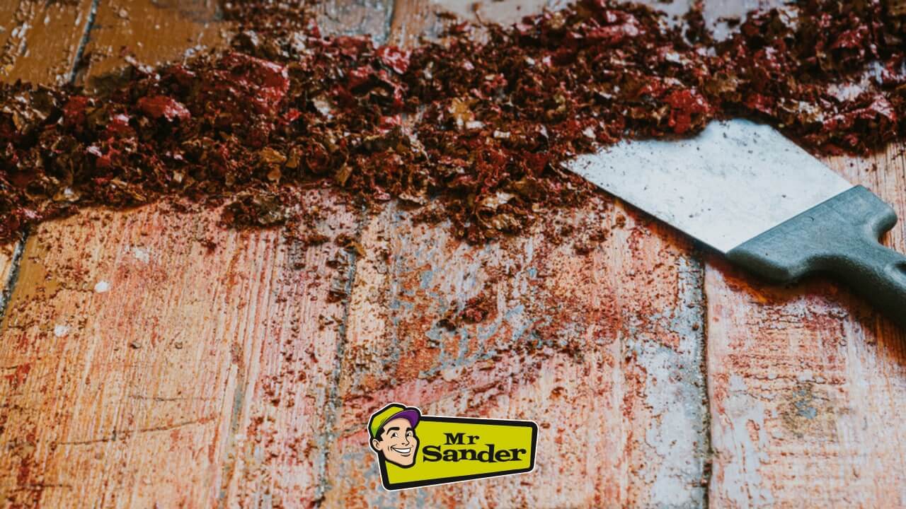 A close-up view of an old wooden floor being scraped clean of paint and debris with a scraper tool. The Mr Sander® logo is displayed.
