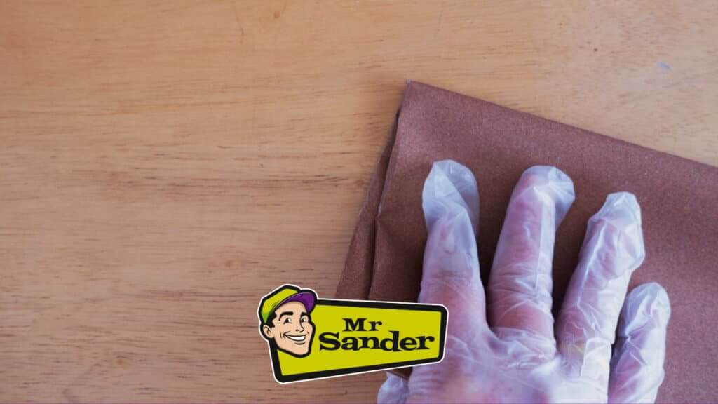 A hand wearing protective gloves holding a piece of sandpaper, working on a wooden surface as part of the floor refinishing process by Mr Sander®.
