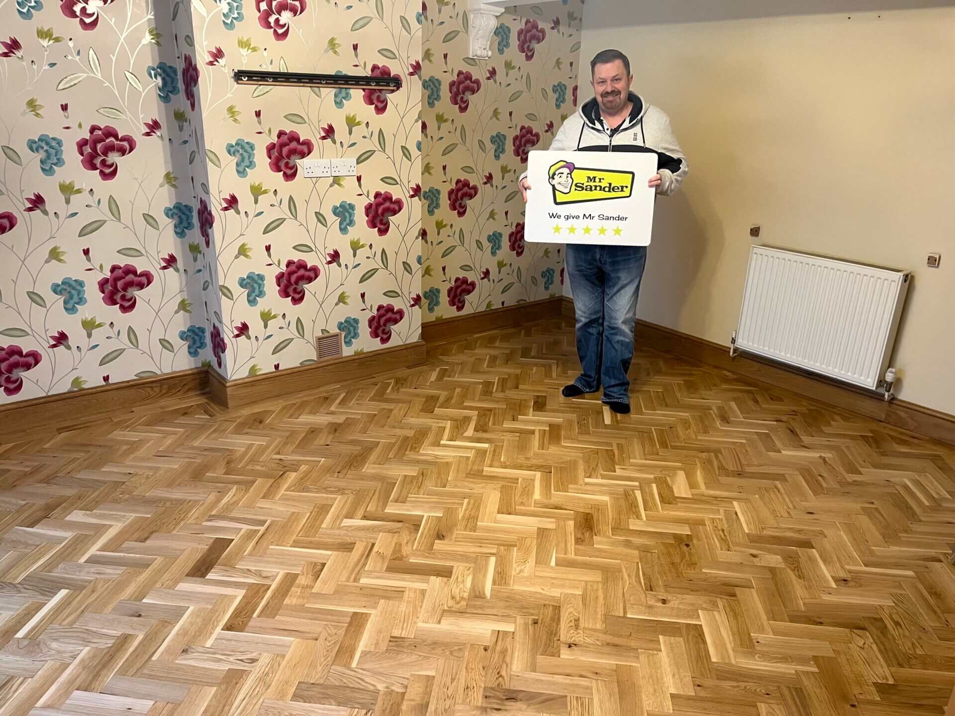 A man holding a Mr Sander® sign stands in a room with a beautifully restored oak herringbone parquet floor, showcasing the refinished flooring.