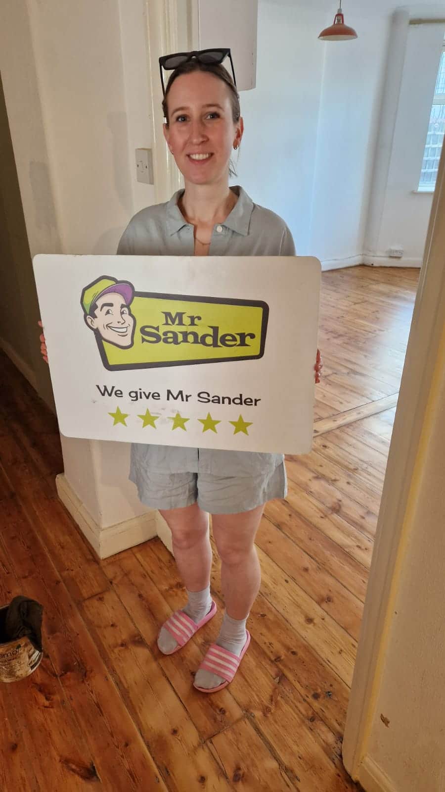 A happy customer standing in a room with newly refinished hardwood floors in West London, UK, holding a sign that reads "Mr Sander® - We give Mr Sander 5 stars". The room features natural light and simple, elegant decor.