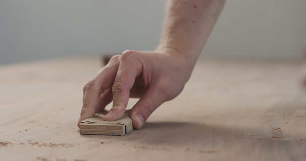 A close-up of a hand using sandpaper on a wooden surface, illustrating the precision involved in Hand Sanding vs. Machine Sanding.