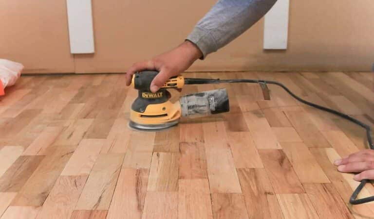 Hand holding a yellow and black orbital sander on a hardwood floor.