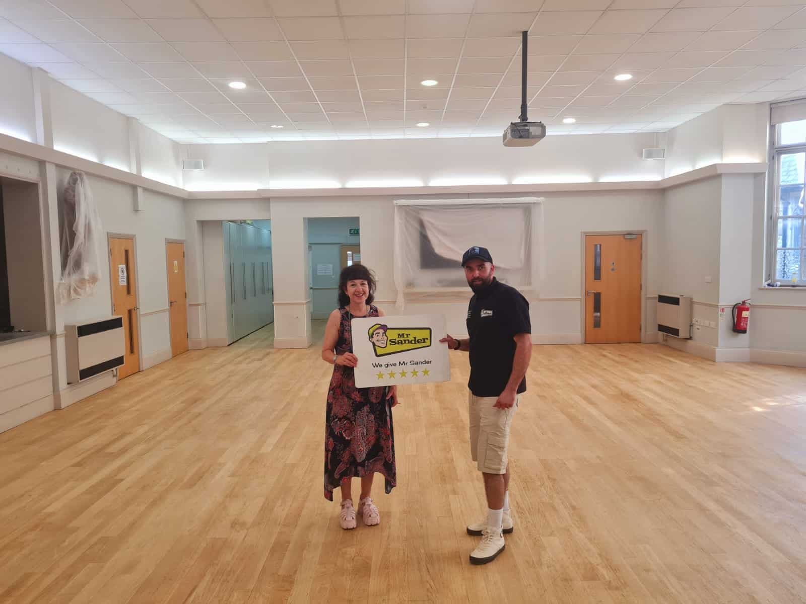 A satisfied customer standing with a Mr Sander® technician in a newly sanded and refinished commercial space. The customer holds a sign with the Mr Sander® logo and a five-star rating, showcasing the excellent workmanship.