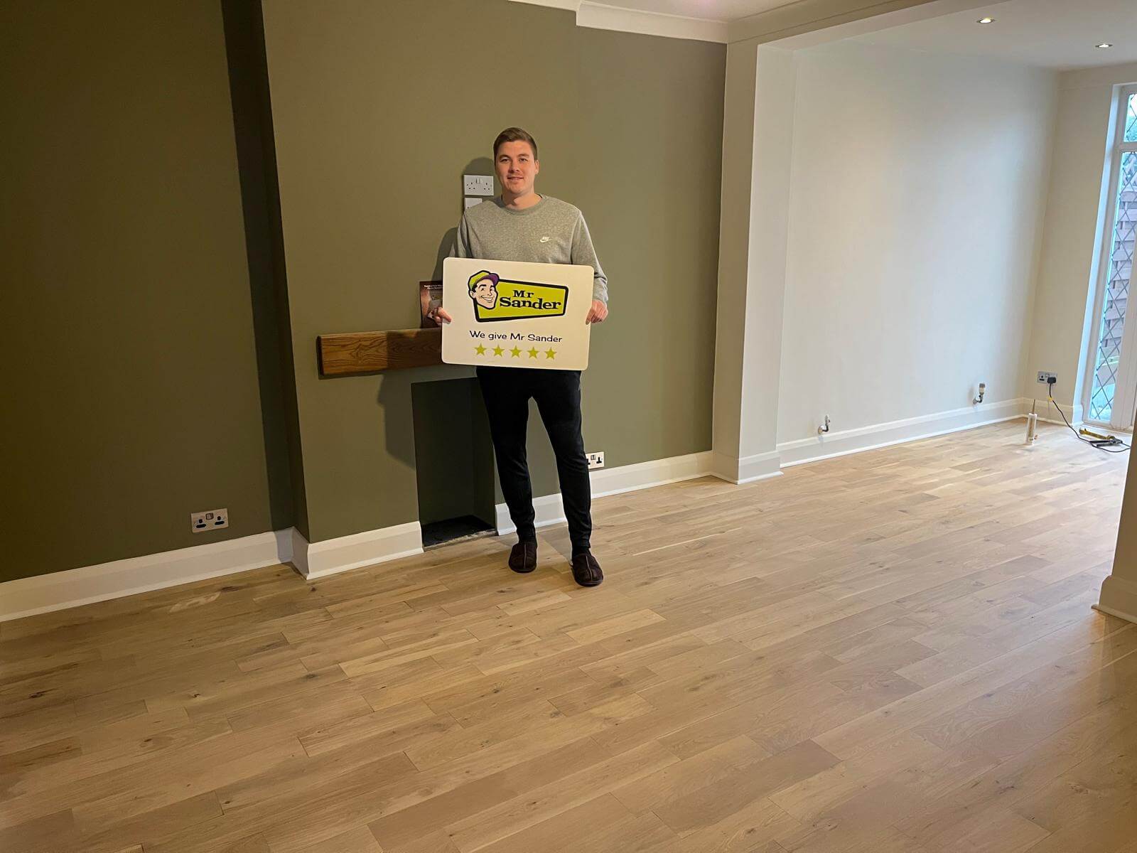 A happy customer holding a sign with a 5-star rating for Mr Sander, showcasing a beautifully fitted wooden floor in their UK home.
